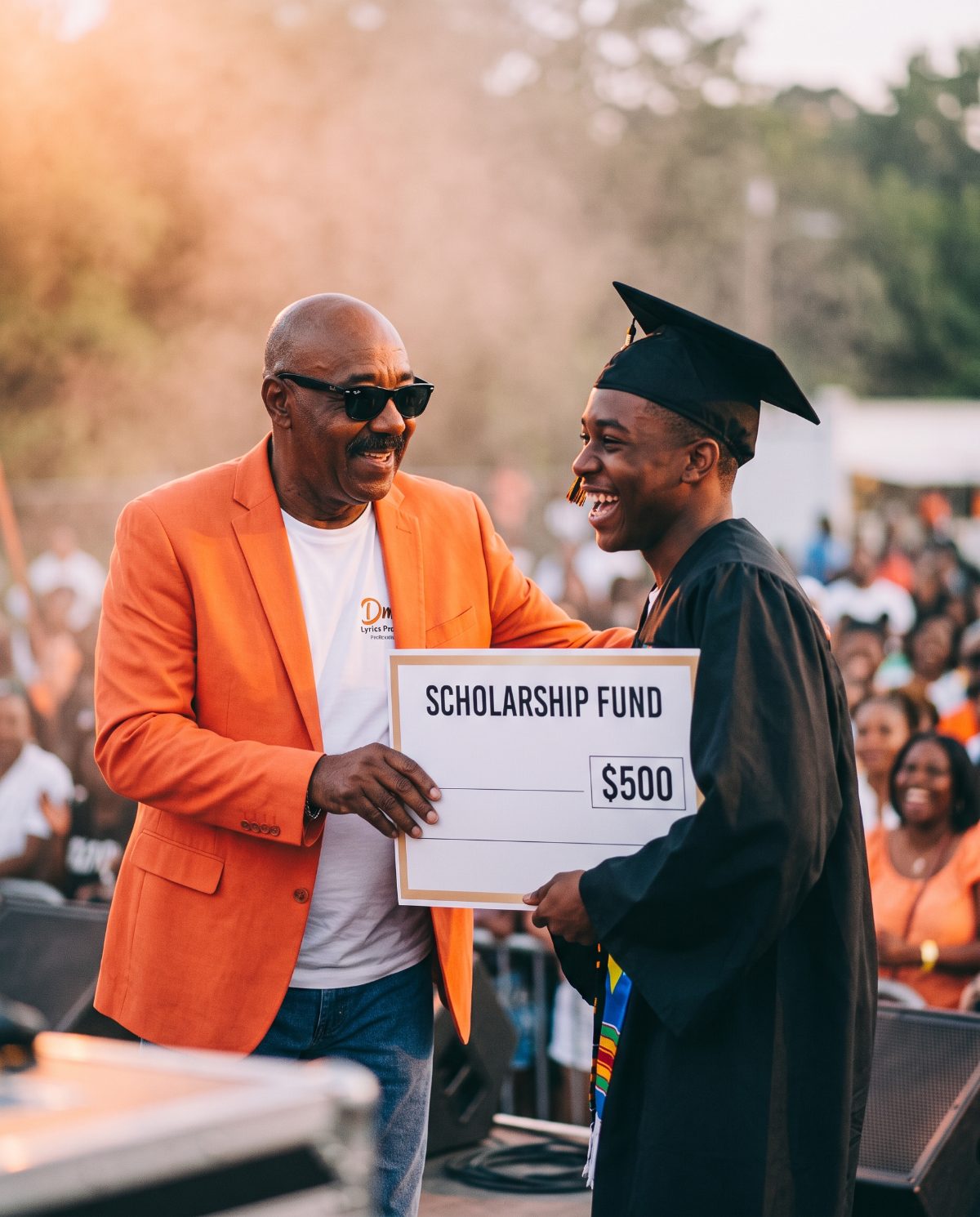 DMANLYRICS in his orange jacket presenting a $500 Scholarship Fund cheque to a smiling graduate
